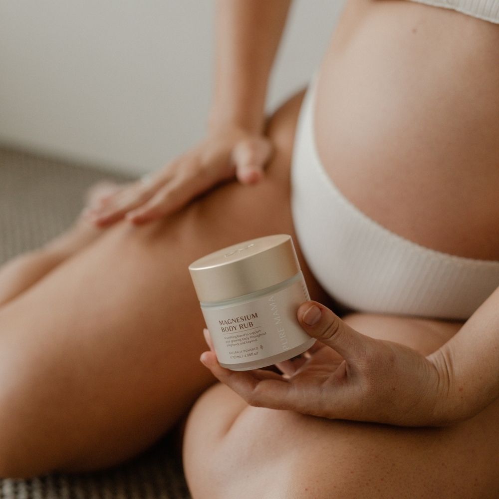 A person in white underwear sits on the floor applying Pure Mama Magnesium Body Rub to their thigh for muscle relaxation, highlighting the product jar and their hands.
