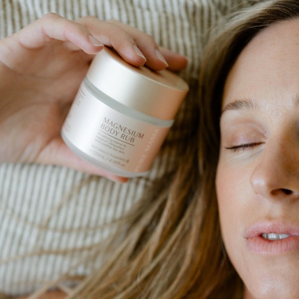 A woman with closed eyes lies on a striped surface, holding a Pure Mama Magnesium Body Rub jar with SyriCalm™ near her face, highlighting its soothing pain relief effects.